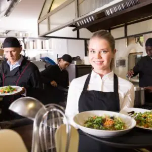 Smiling waitress serving salad in restaurant kitchen.
