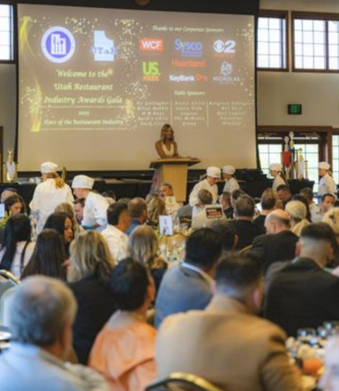 A speaker addresses a large audience at a formal event in a bright venue.